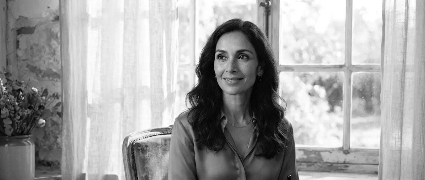 A woman seated by a sunlit window in a soft pink blouse